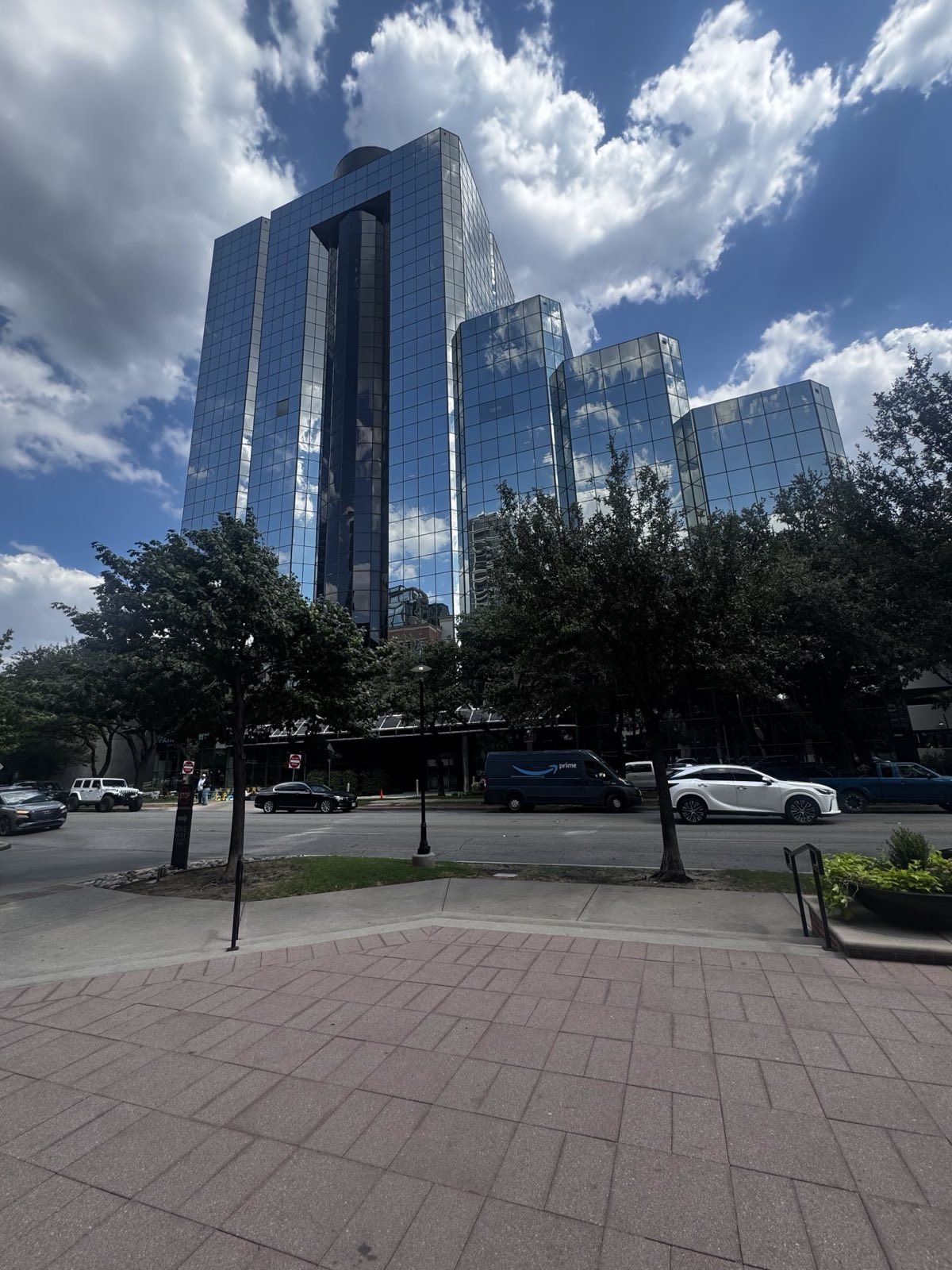 Street view of Highland Park Place at 4514 Cole Avenue, a mirrored glass office tower rising above shade trees, parked cars and the sidewalk out front